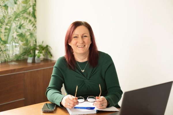 Lucy Dawson sits at a desk holding her glasses away from her face. She is smiling at the camera, creating a warm and approachable feel. A laptop, notebook, pen and phone sit on her desk.